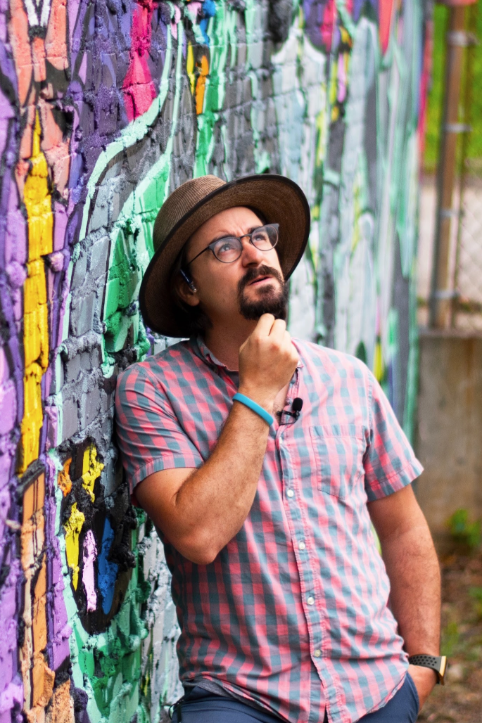 A man with glasses, a beard, and a mustache is wearing a wide-brimmed hat, a pink and blue checkered shirt, and a blue wristband. He is standing and leaning against a brick wall with colorful graffiti, looking up thoughtfully with his hand on his chin.