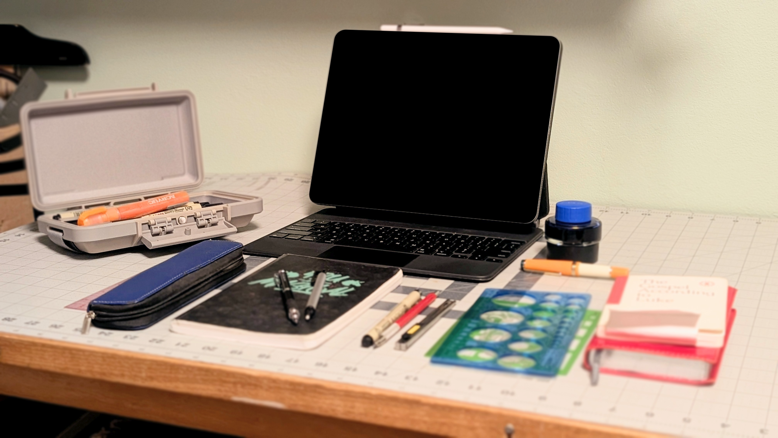 A cluttered desk with a closed laptop, a blue pencil case, a notebook with pens, a ruler, a pen case, a small ink bottle, two books, and a circle guides.