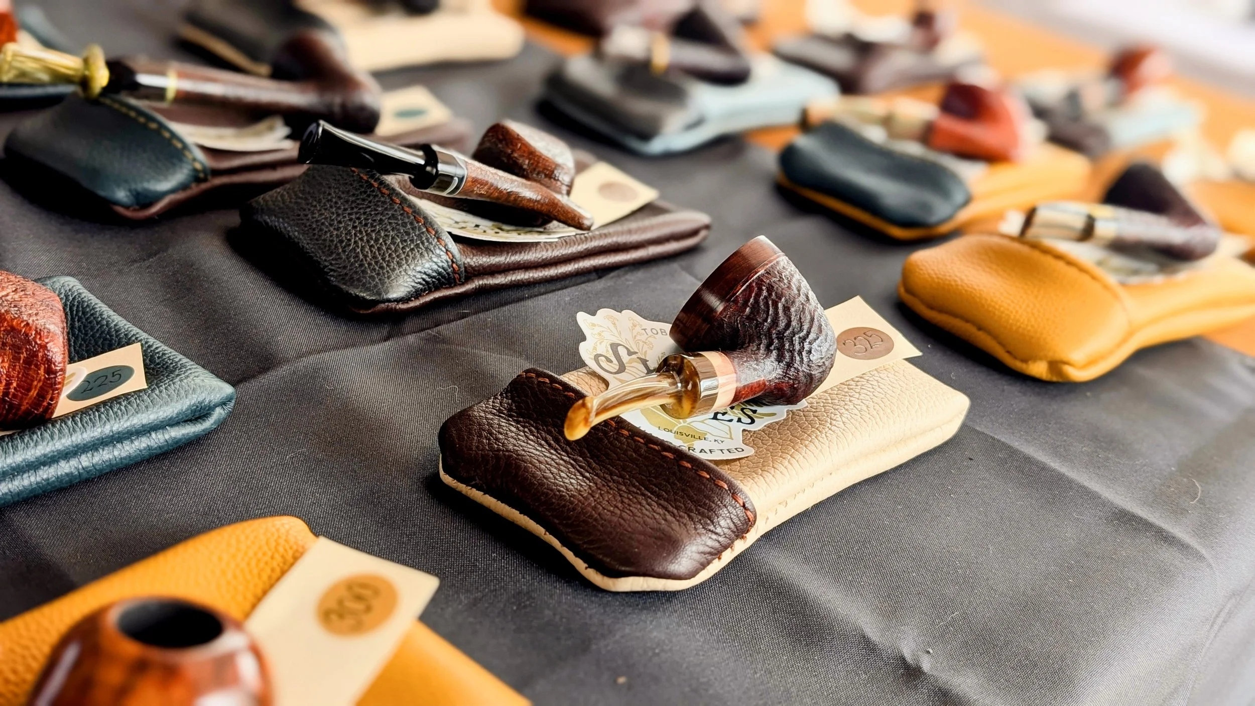 Small leather bag to store pipes and several smoking pipes displayed on a table at a market or shop.