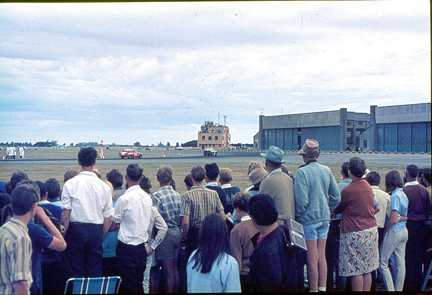 A couple of Anglias pound past a crowd of spectators, with only a strip of grass and a rope between them