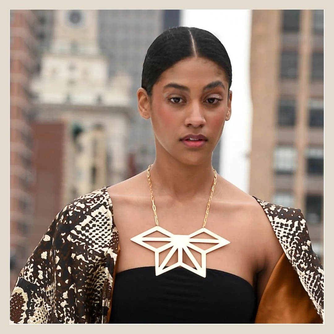 Asanoha hemp leaf statement necklace styled on a model in a middle part, wearing a strapless black top, a patterned coat, and a large geometric white necklace, standing outdoors with a cityscape of tall buildings in the background.