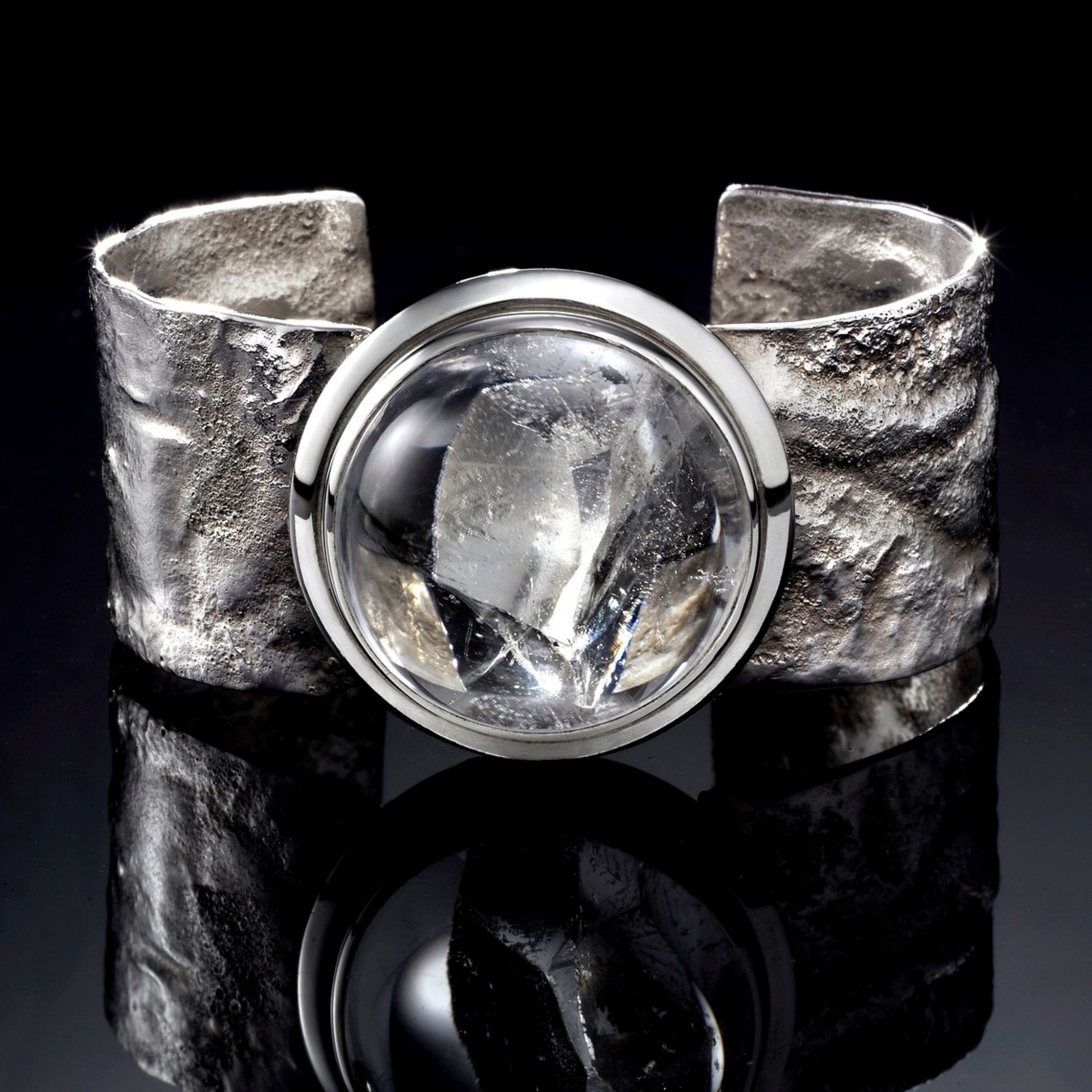 An artisan hand-forged sterling silver cuff bracelet with a wide, rustic reticulated silver band and a prominent bezel-set round clear quartz crystal with internal phantom inclusions