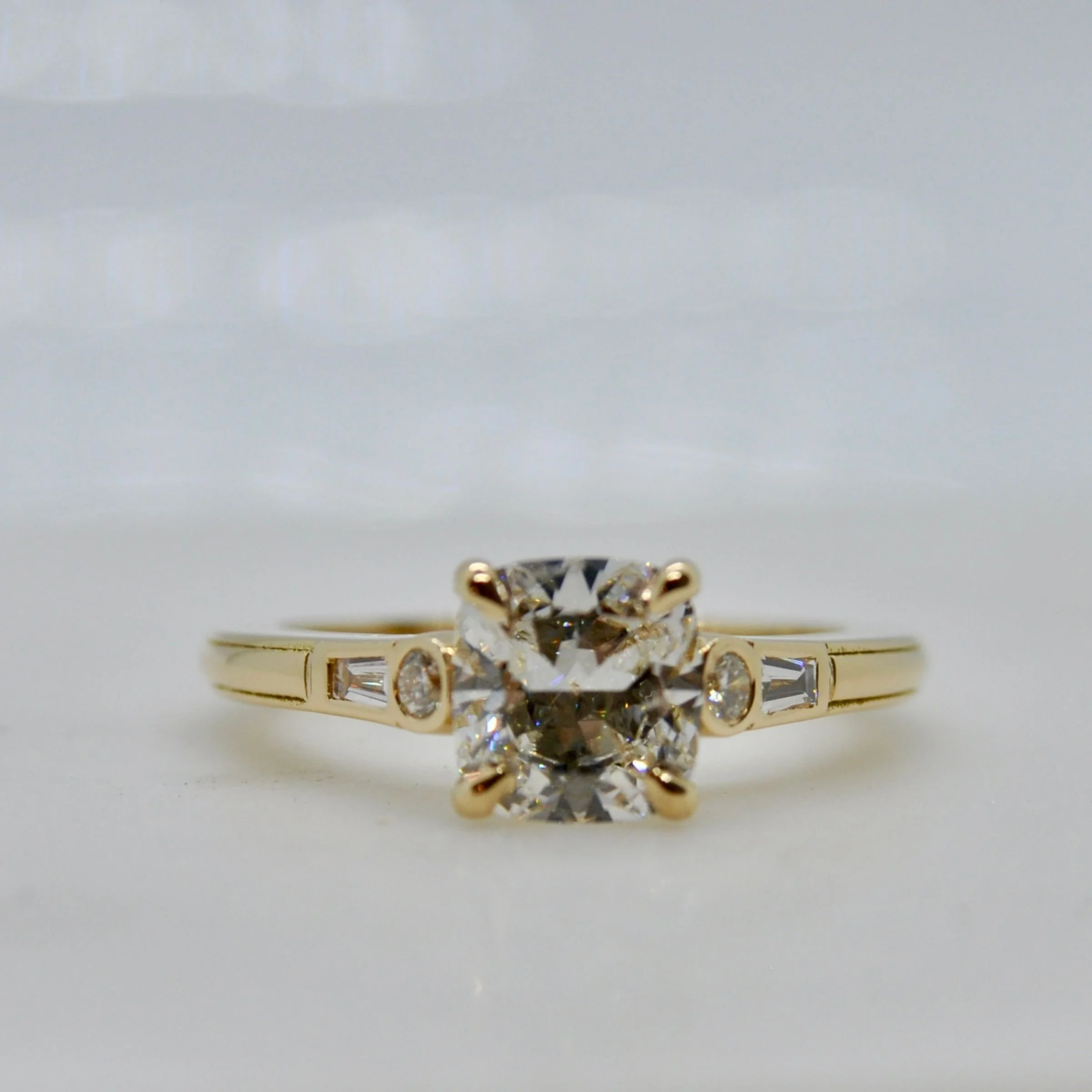 A modern three-stone 18k yellow gold engagement ring showcasing a brilliant cushion-cut diamond center stone with small round accents and baguette side diamonds on a sleek polished band