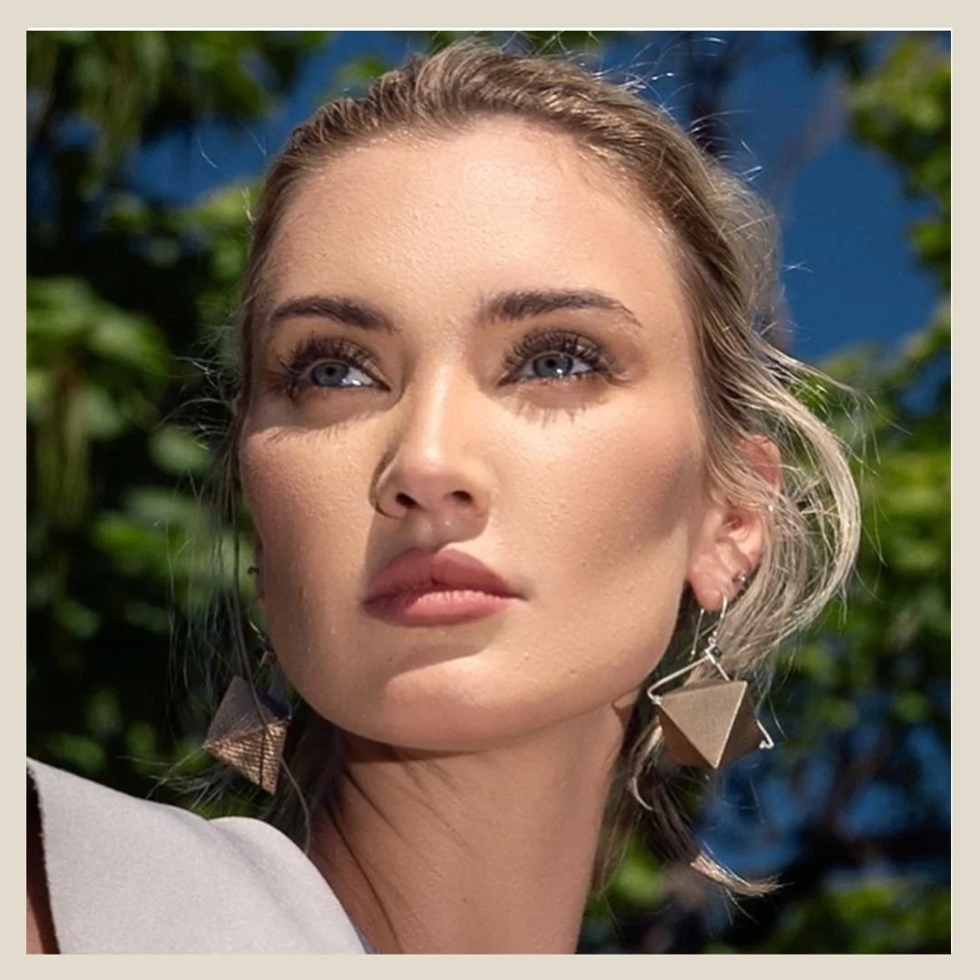 Model wearing 3d triangle dangle earrings. Close-up of a young woman with blonde hair, blue eyes, wearing geometric earrings, outdoors with green leaves and a blue sky in the background.
