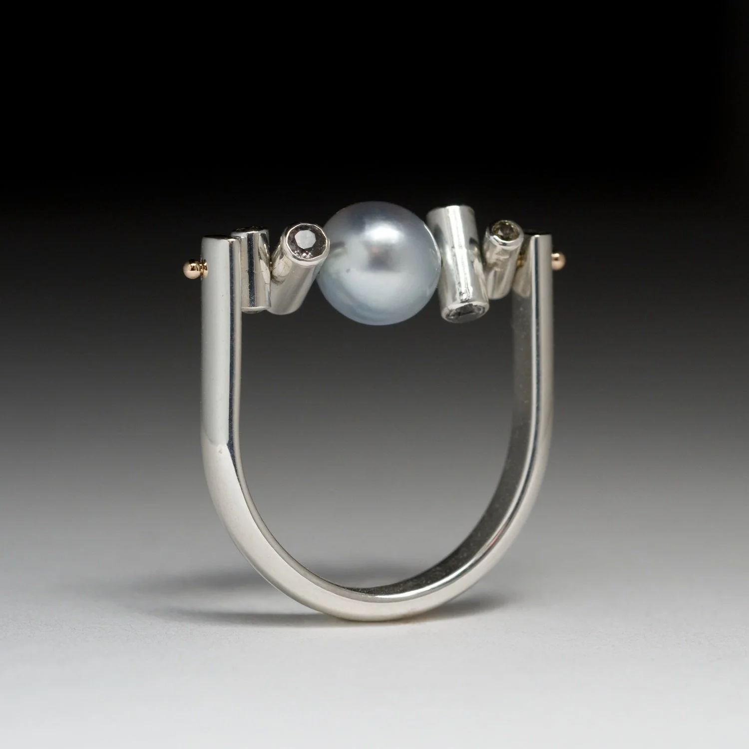 An artisan modern silver statement ring showcasing an industrial U-shaped band with an off-center blue-grey Tahitian pearl, small tube details with green sapphires, yellow gold pins for movement.