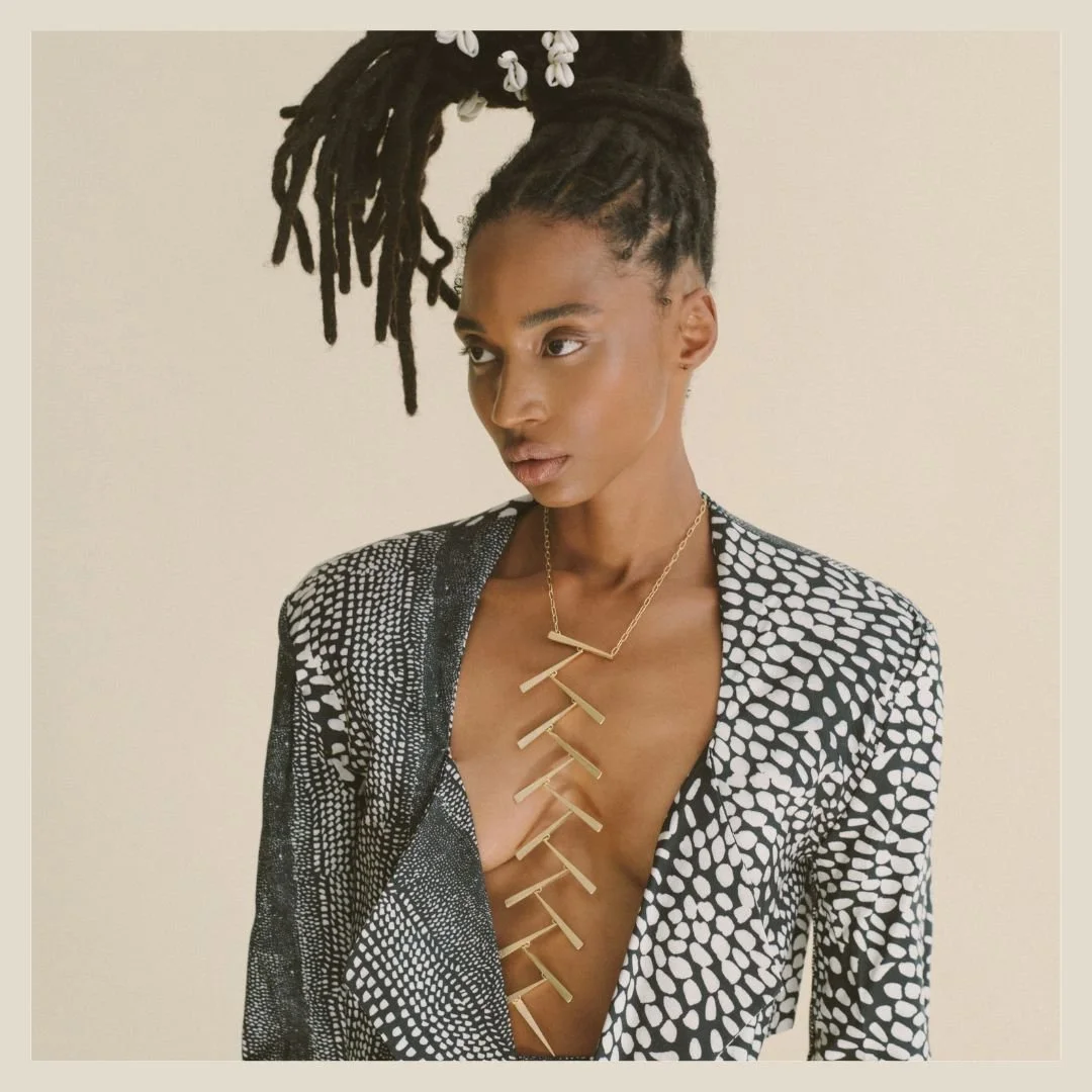 A woman with dark skin, has a long, gold, geometric necklace extending down her chest. Intricate dreadlocks styled in a high bun with decorative white beads, is wearing a black and white patterned blazer with a plunging neckline.
