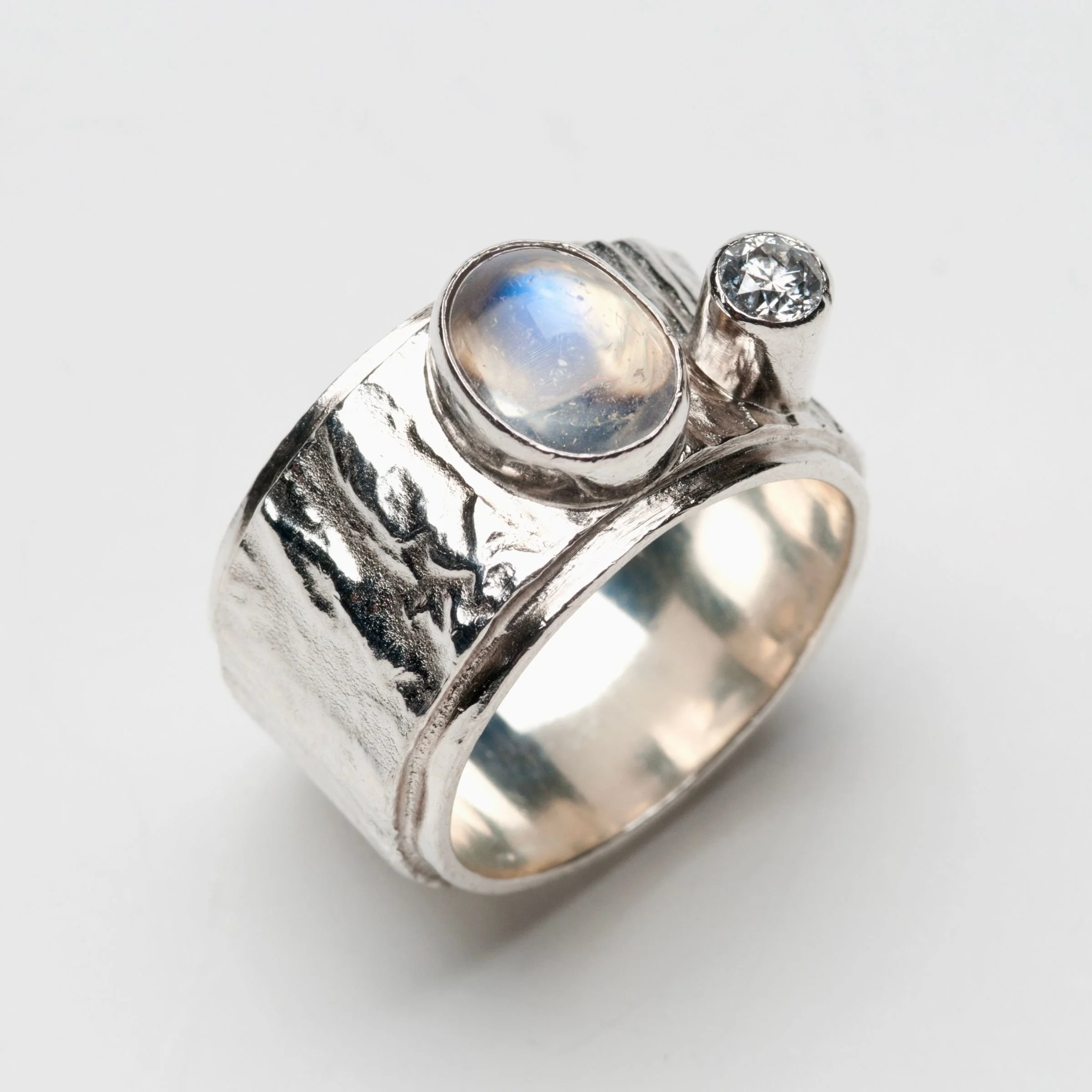 wide, reticulated textured sterling silver band ring featuring a prominent oval cabochon moonstone with a blue sheen and a smaller, bezel set round diamond accent stone.