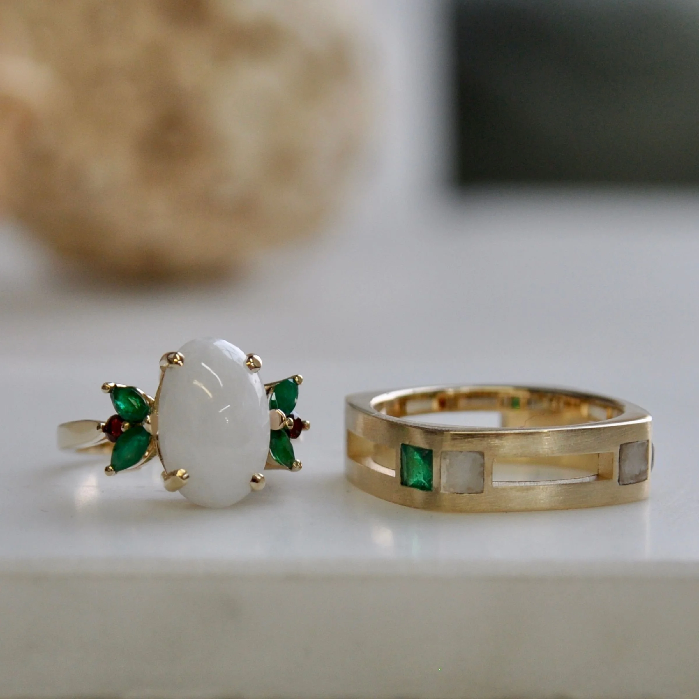 14k yellow gold wedding band featuring an open-channel geometric design inset with a square-cut green emerald and a small inset marble next to a matching engagement ring with a large oval white cabochon green emeralds and round red garnets