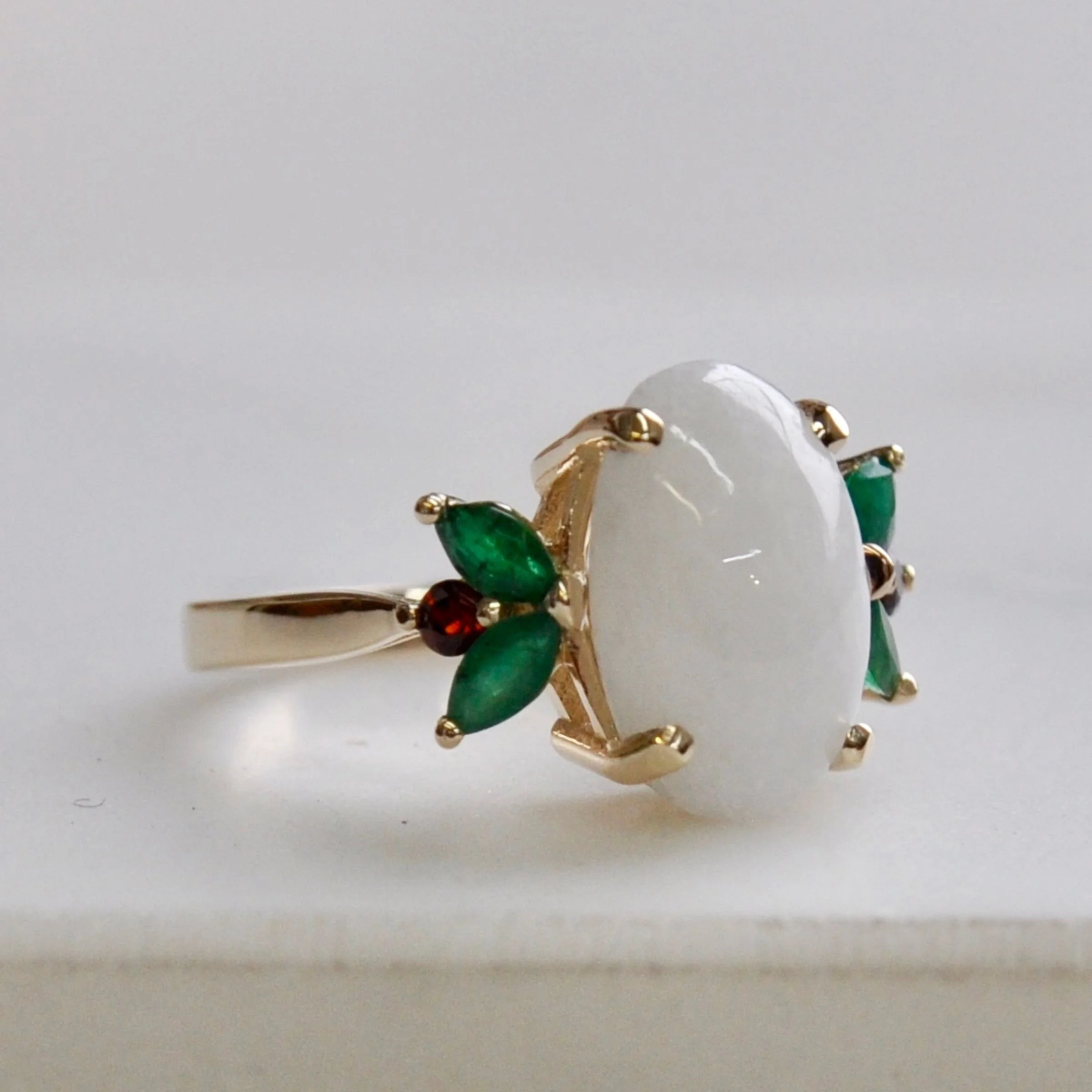 A custom 14k gold engagement ring featuring a large oval opal cabochon flanked by marquise-cut green emerald side stones and round red garnets arranged in a floral pattern.