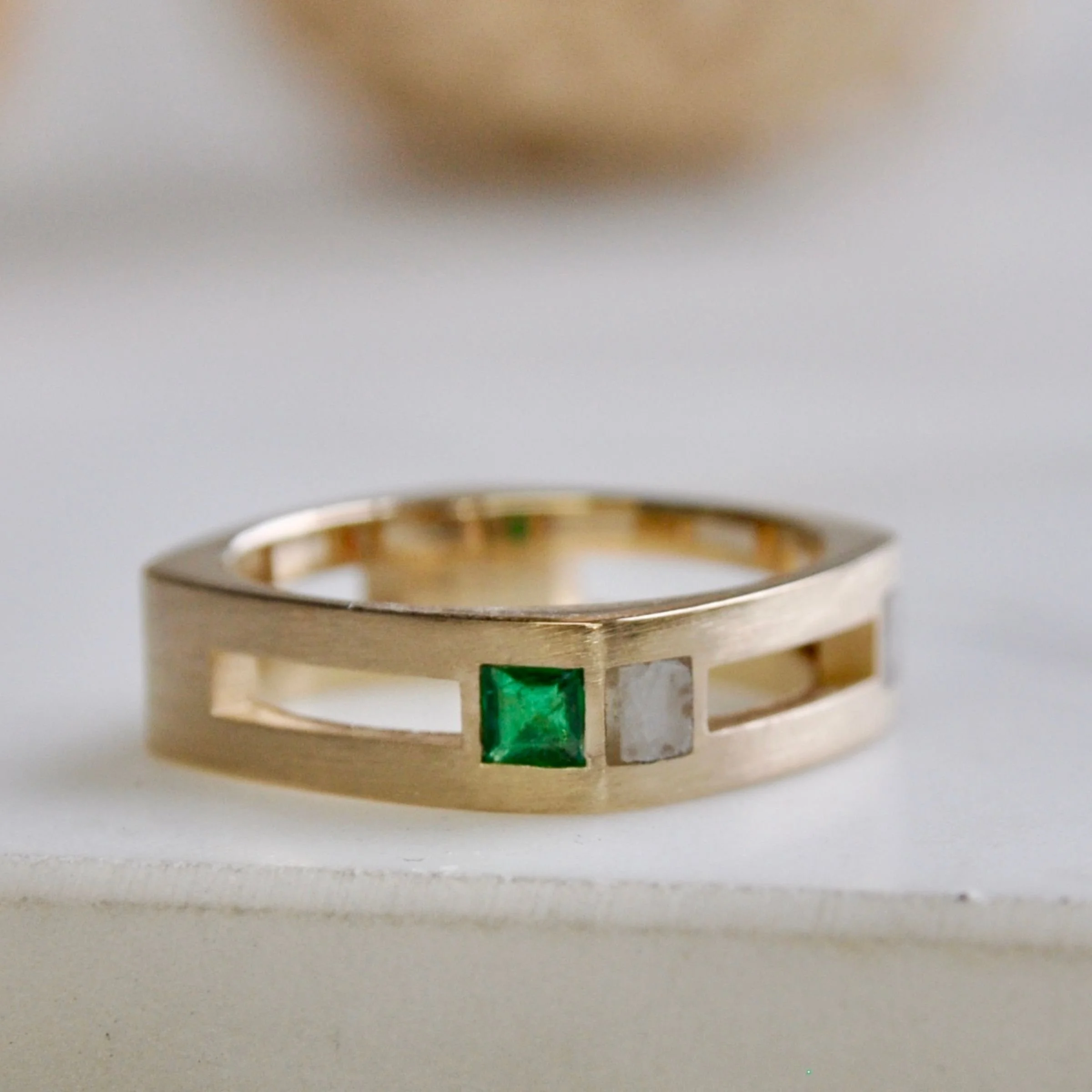 A custom modern wedding band 14k yellow gold ring featuring an open-channel geometric band inset with a square-cut green emerald and a small found object marble with a matte finish.
