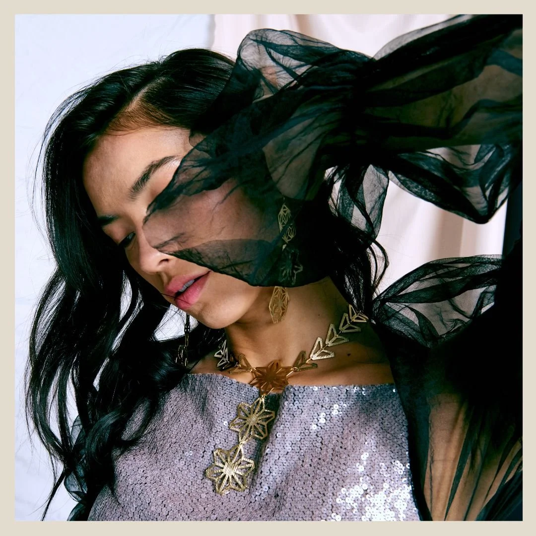A woman wearing a statement necklace patterned asanoha hemp leaf large gold floral design, holding a sheer black-green cloth partially covering her face.
