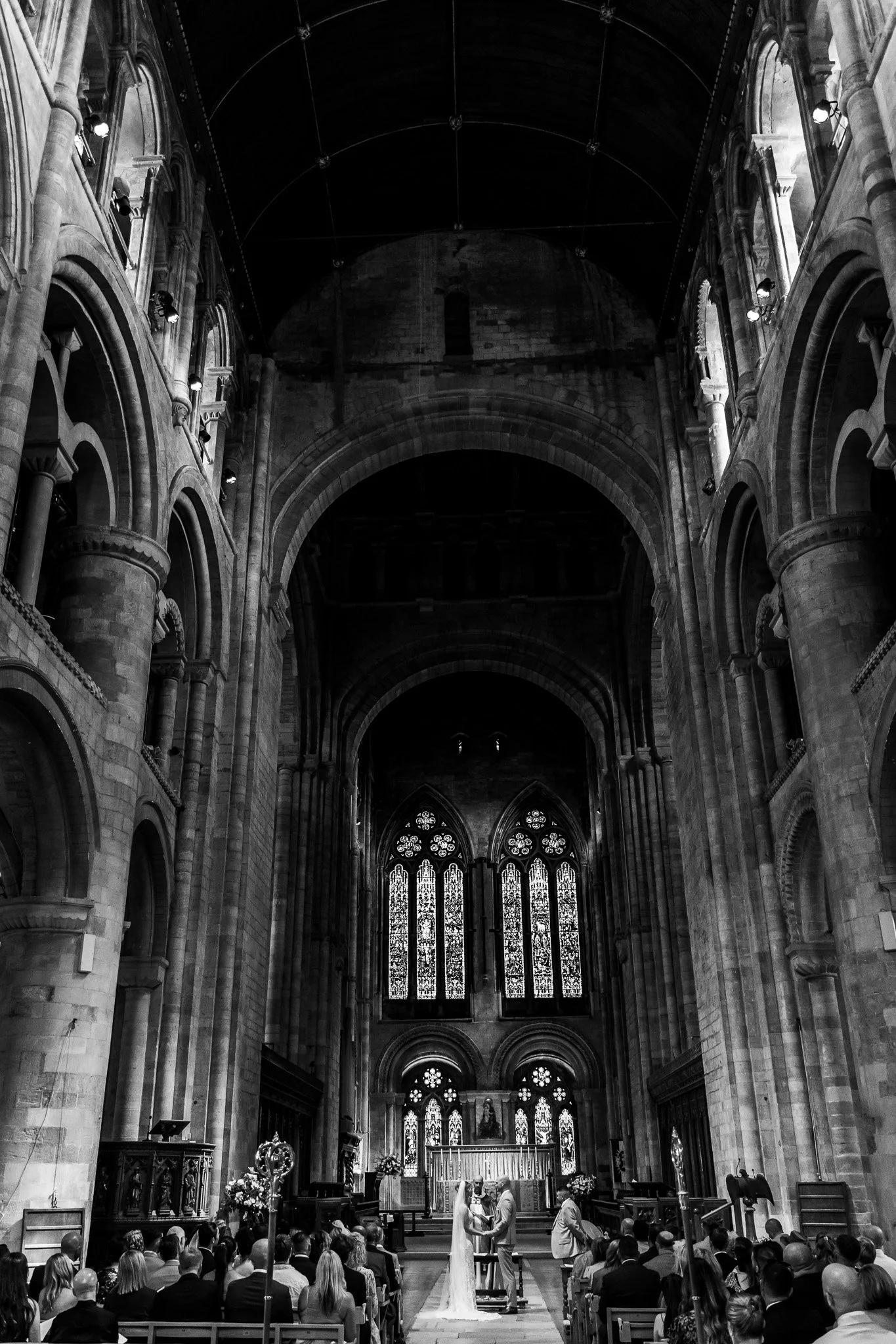 Black and white photo of a wedding ceremony inside a large Gothic cathedral with high vaulted ceilings and stained glass windows. Matthew Johnston Photography, Hampshire, UK.