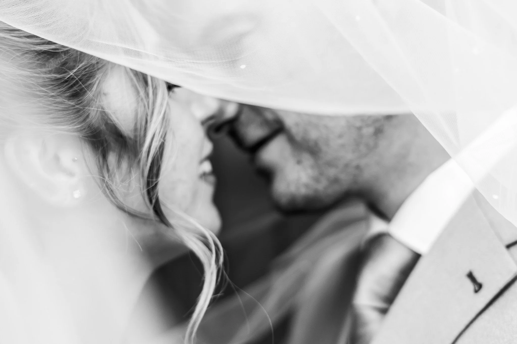 Close-up of a bride and groom about to kiss, with the bride wearing a veil and the groom in a suit, in black and white.
