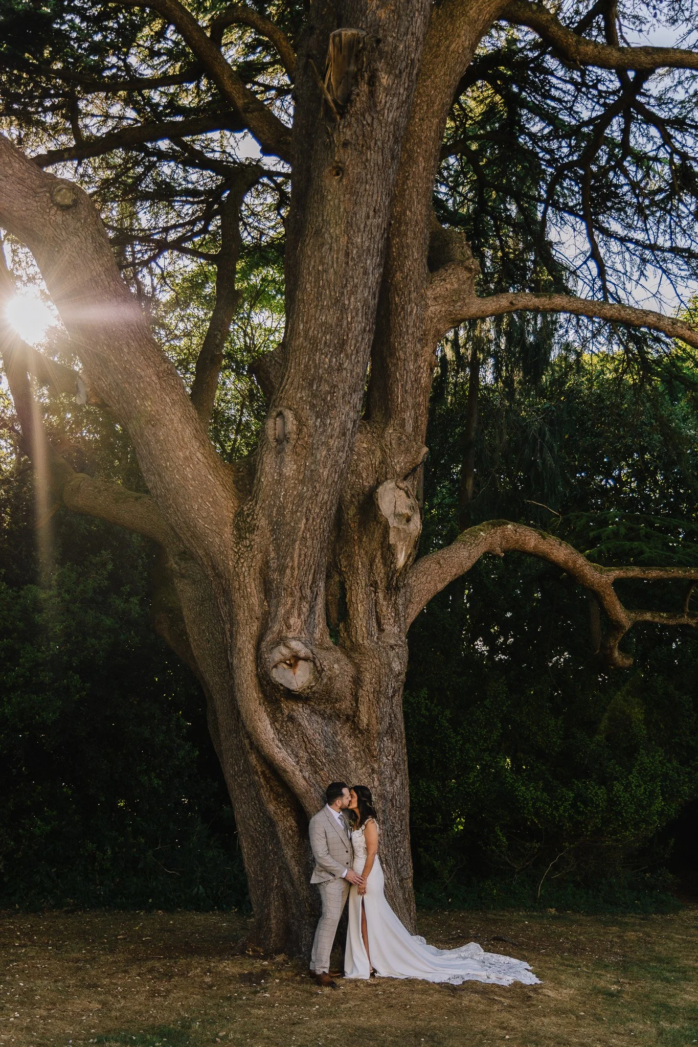 A bride and groom stand close together in front of a large tree, sharing a kiss during their wedding photos. Matthew Johnston Photography, Hampshire, UK.