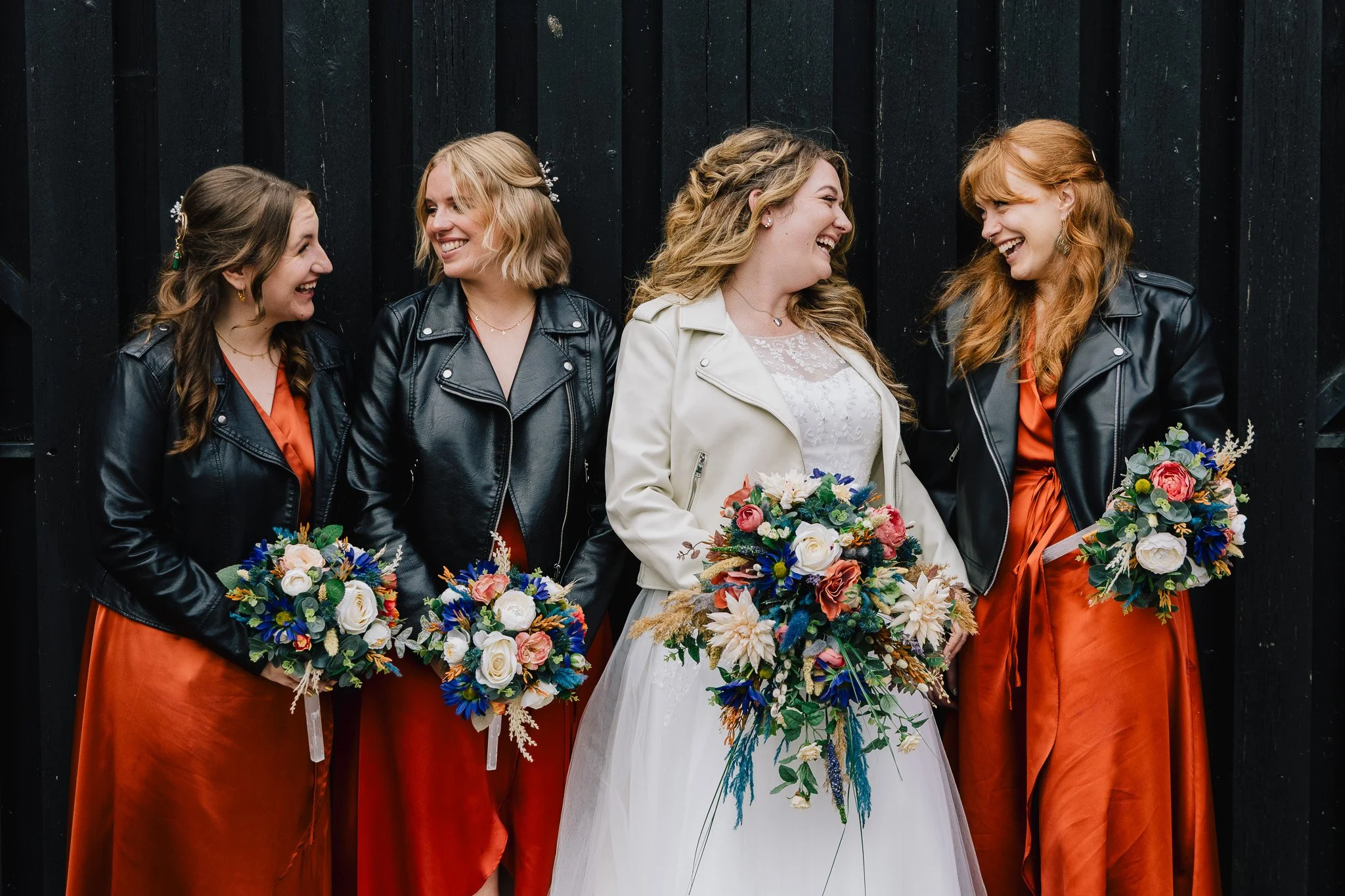 A bride and four bridesmaids, smiling and holding bouquets, dressed in black leather jackets and orange dresses, standing against a black wooden background. Matthew Johnston Photography, Hampshire, UK.