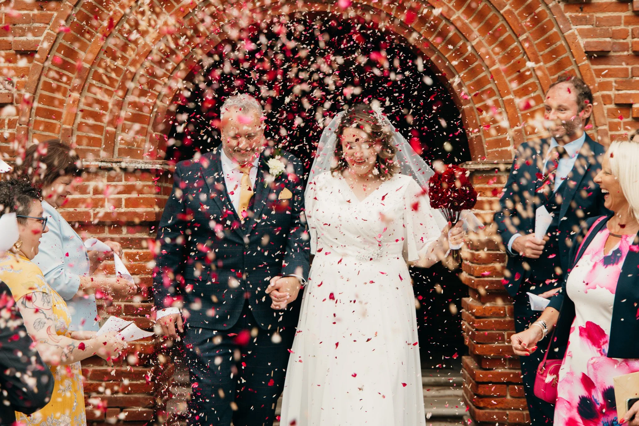 A newlywed couple walking out of a brick archway, holding hands, surrounded by confetti as family and friends celebrate. Matthew Johnston Photography, Hampshire, UK.