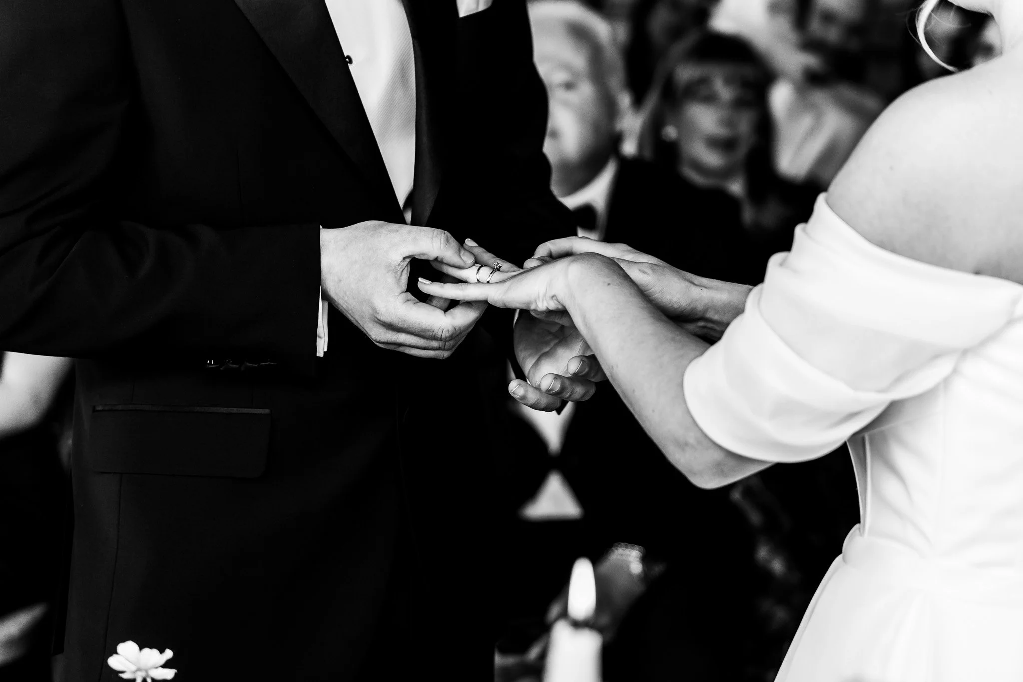 A black and white photo of a couple exchanging wedding rings, with guests in the background.