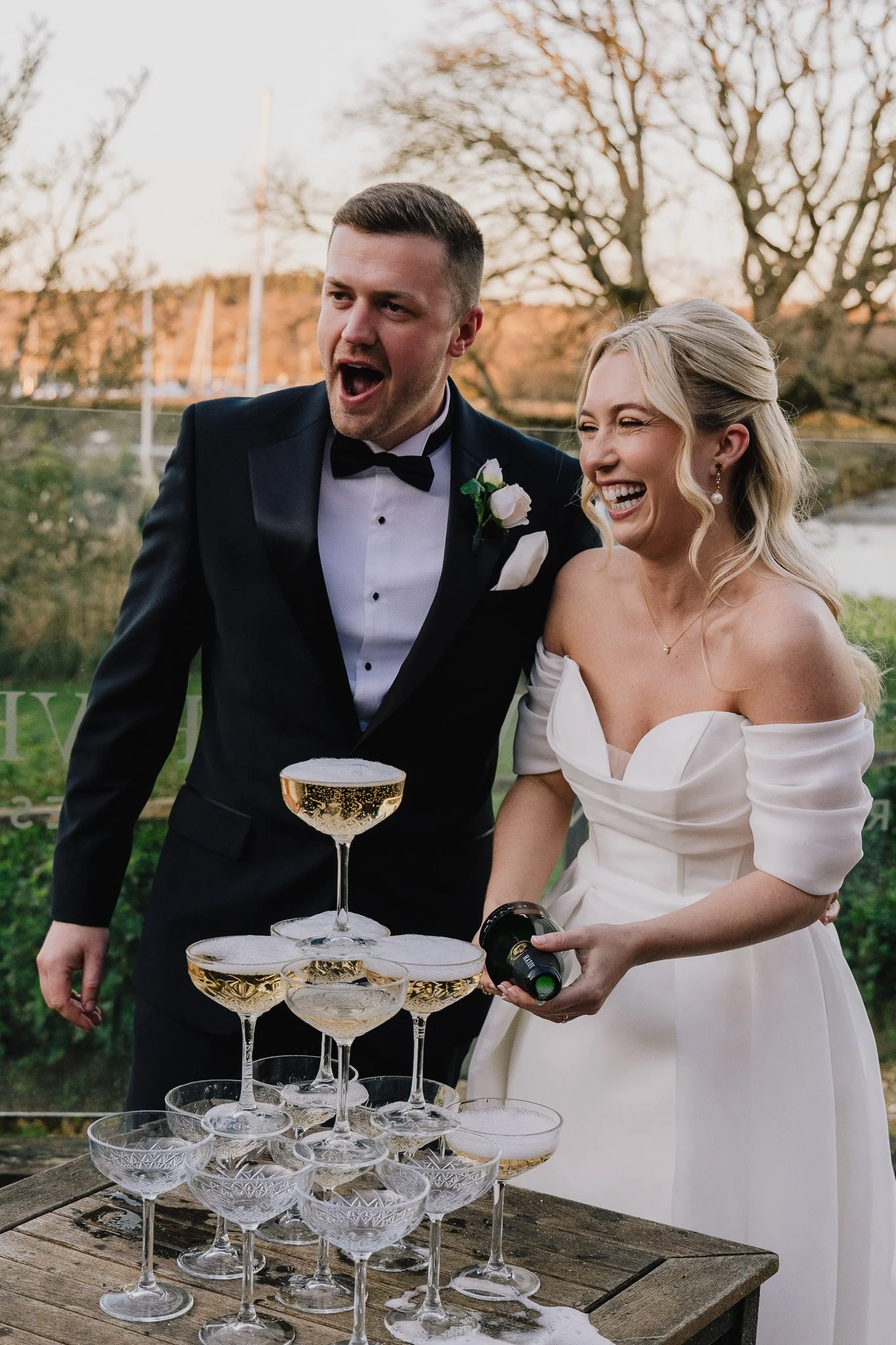 A happy couple on their wedding day, in front of a champagne tower, at The Master Builder’s House Hotel, in Hampshire.