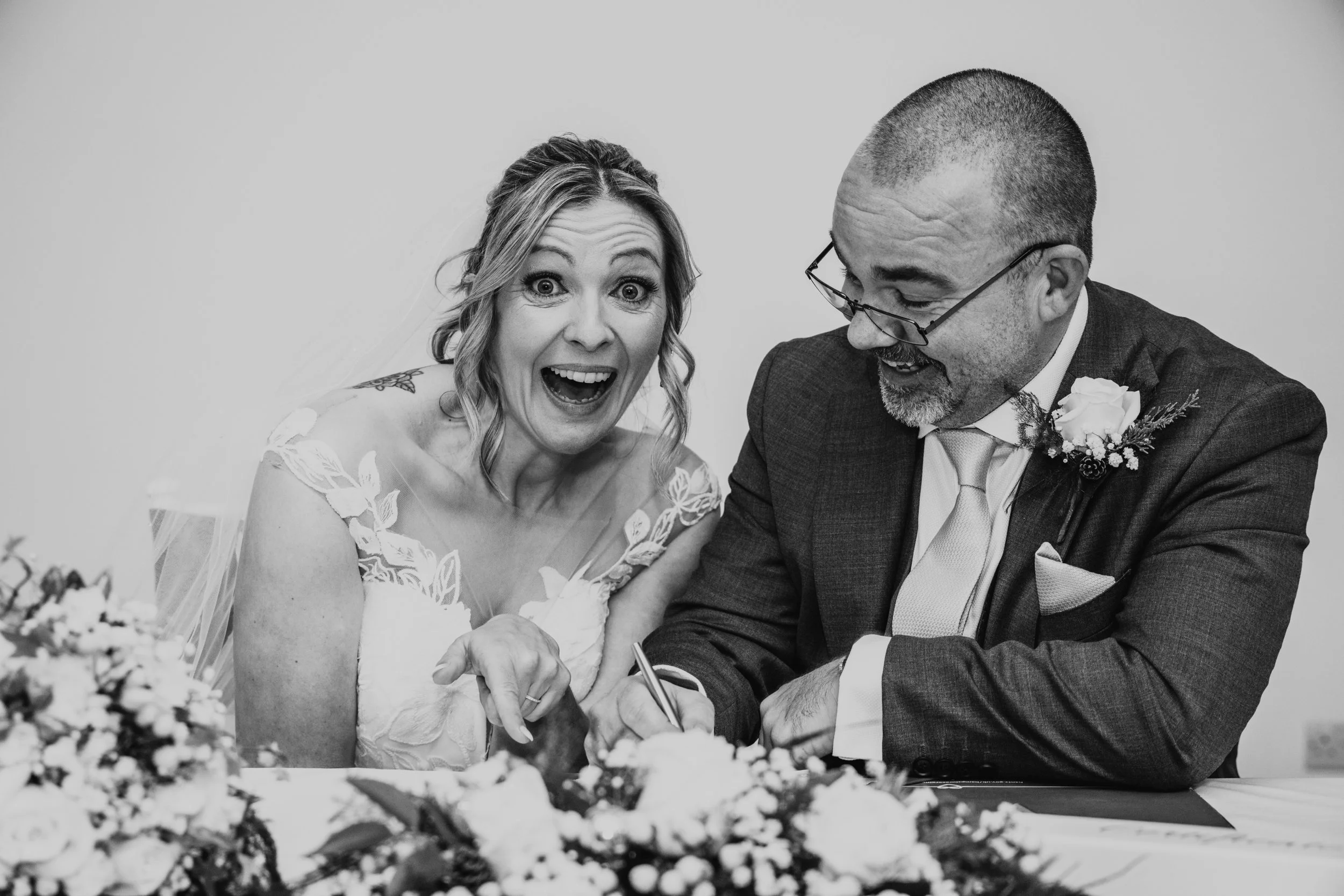 A woman in a wedding dress and a man in a suit are signing a marriage certificate, smiling and celebrating their wedding. Matthew Johnston Photography, Hampshire, UK.