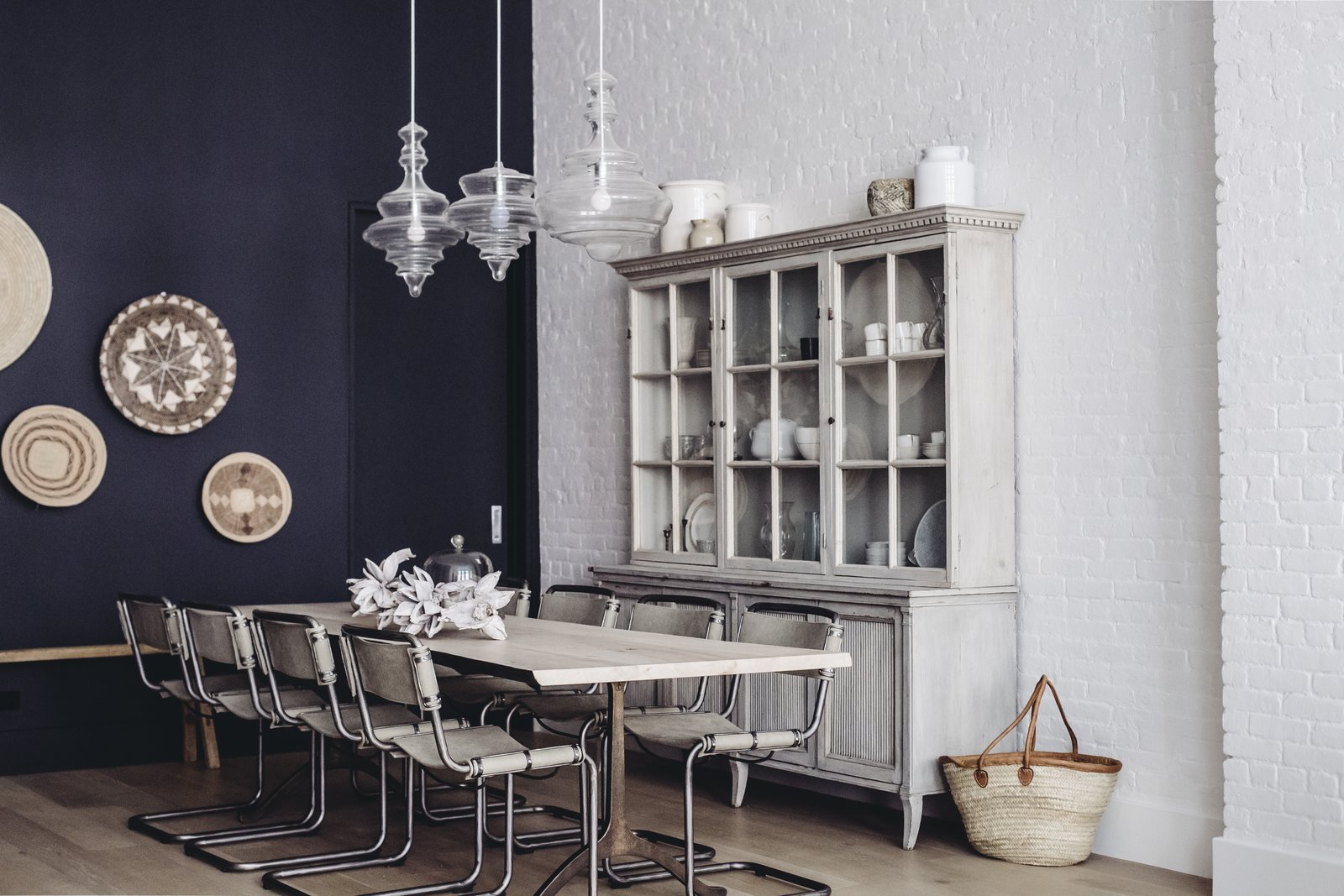 The dining room melds a modern accent wall swathed in "Railings" paint by Farrow and Ball with hanging wall baskets from Habitat Greenwich, bold pendants by Hudson Valley Lighting, and an antique hutch from Lone Ranger Antiques. In the center sits a dining table, custom created with Studio Endo, and chairs from Sarreid. (Chloe Leroux)