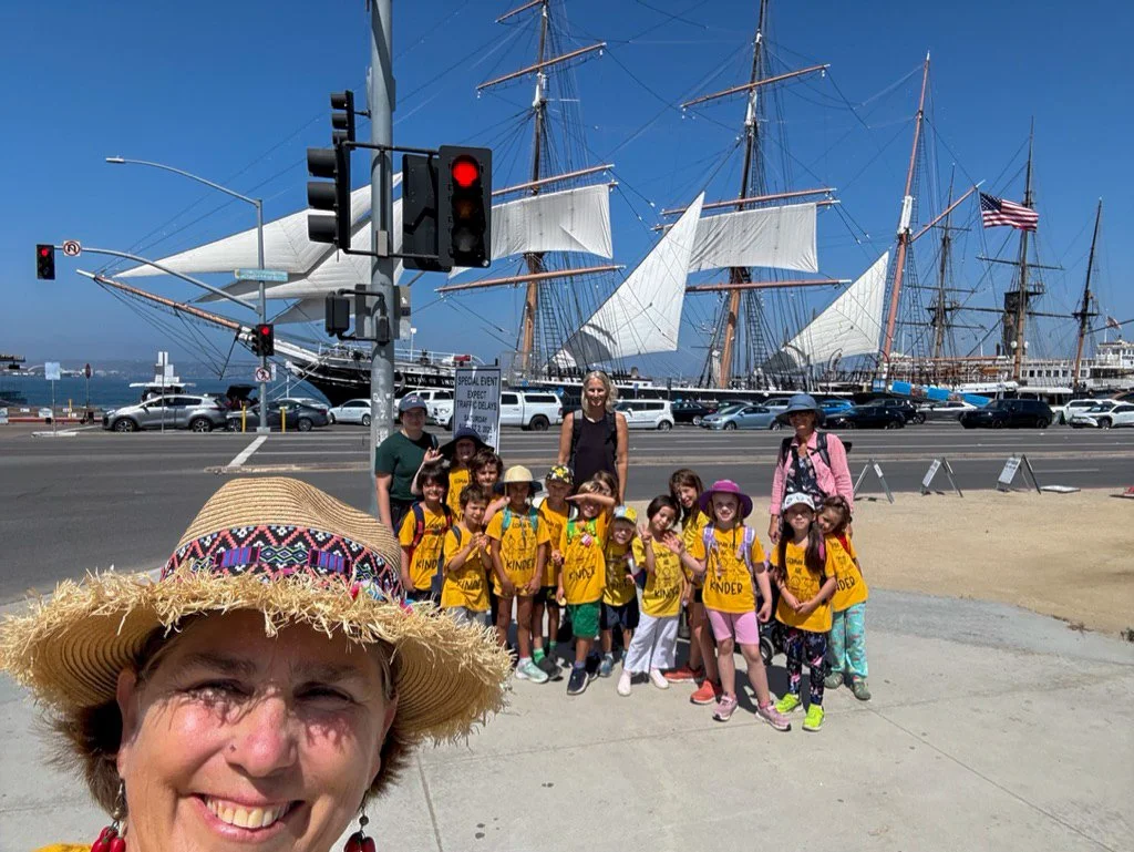 Group of summer camp participants posing in front of a tall ship docked at a harbor, with a teacher smiling in front of the camera.