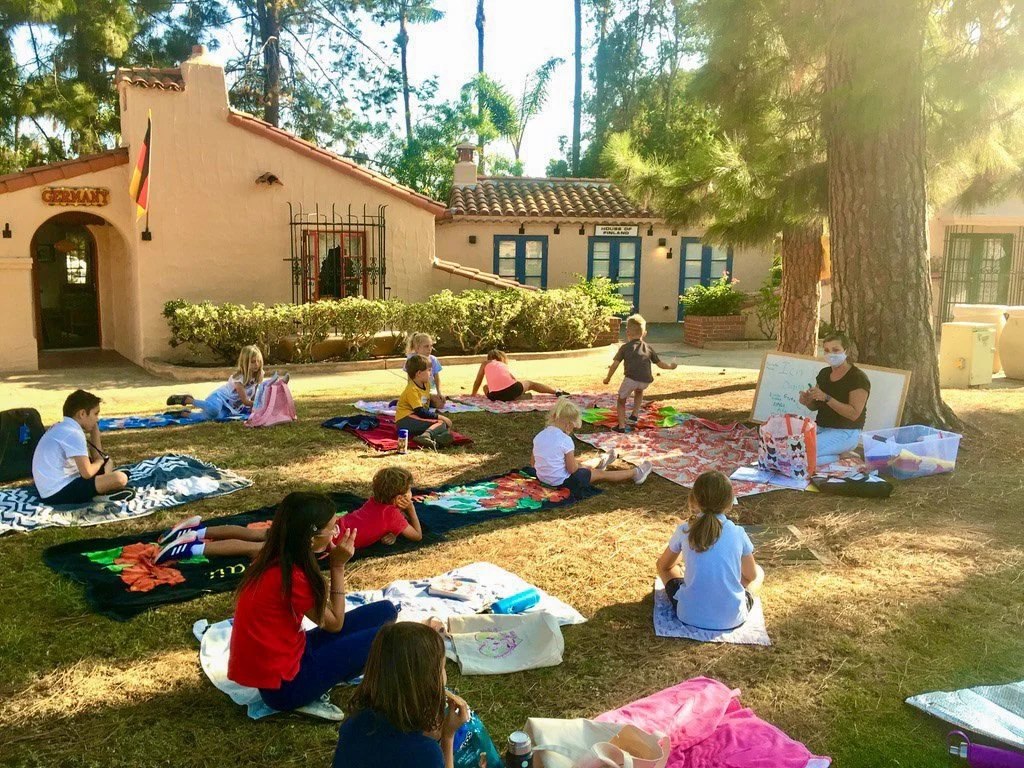 Kinderwerkstatt students sitting outside of the House of Germany cottage during Covid times, listening to their teacher.