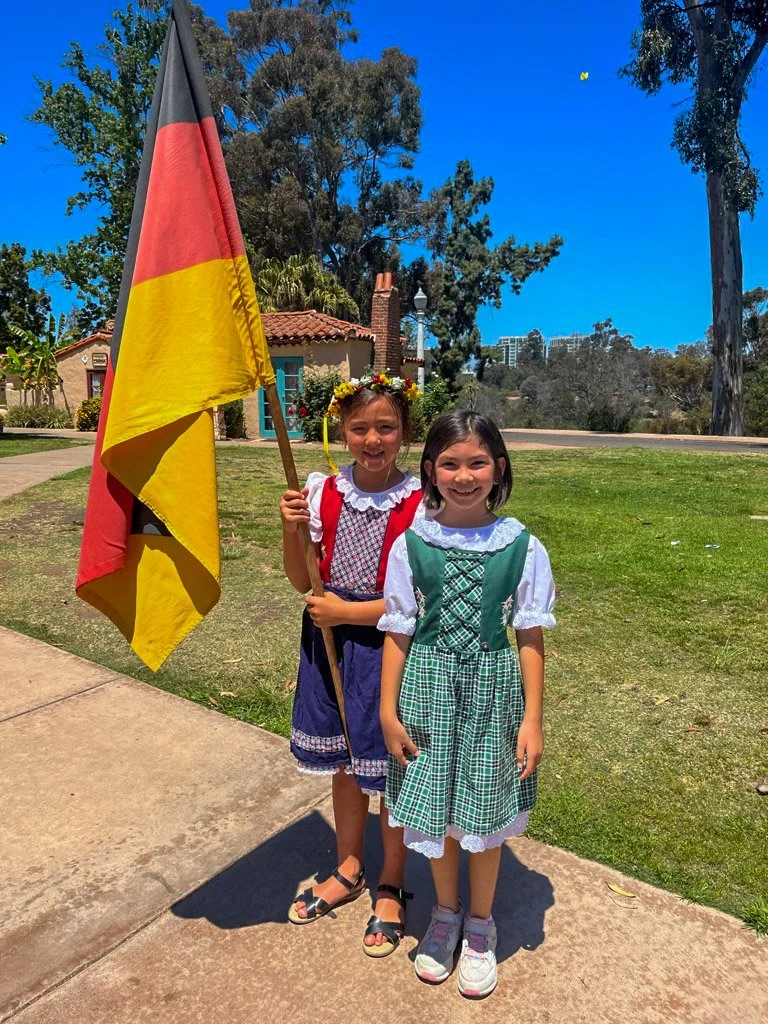 Two young girls dressed in traditional German costumes near the International Cottages, holding an German flag.