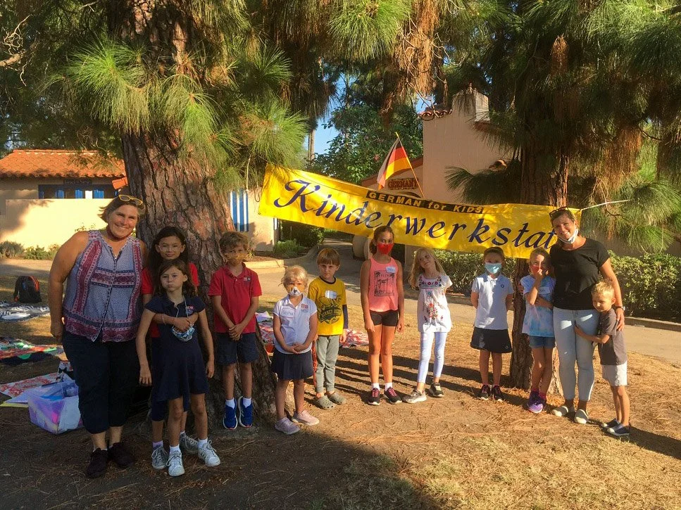 Group of Kinderwerkstatt students posing with their teachers in front of Kinderwerkstatt banner, with some of them wearing masks.