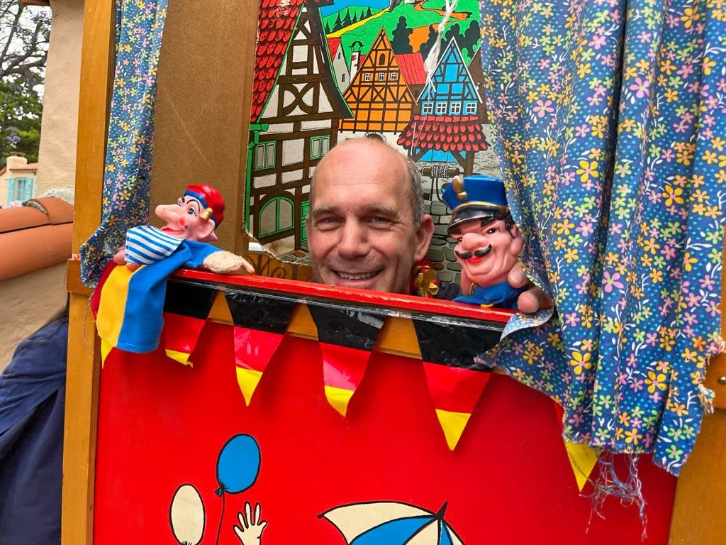 A man smiling behind a puppet theater holding two puppets.