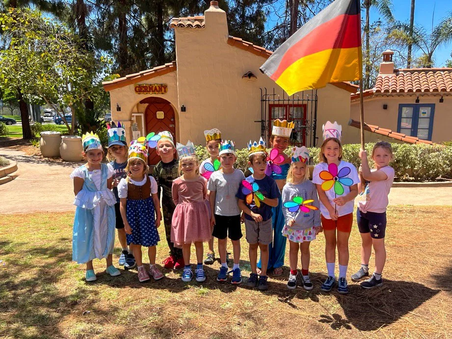 Kinderwerkstatt students dressed in costumes and wearing crowns, holding colorful pinwheels, gathered outdoors under a large German flag, with the House of Germany in the background.