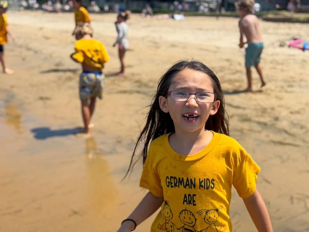 Young summer camp participant standing on a sandy beach surrounded by other children.