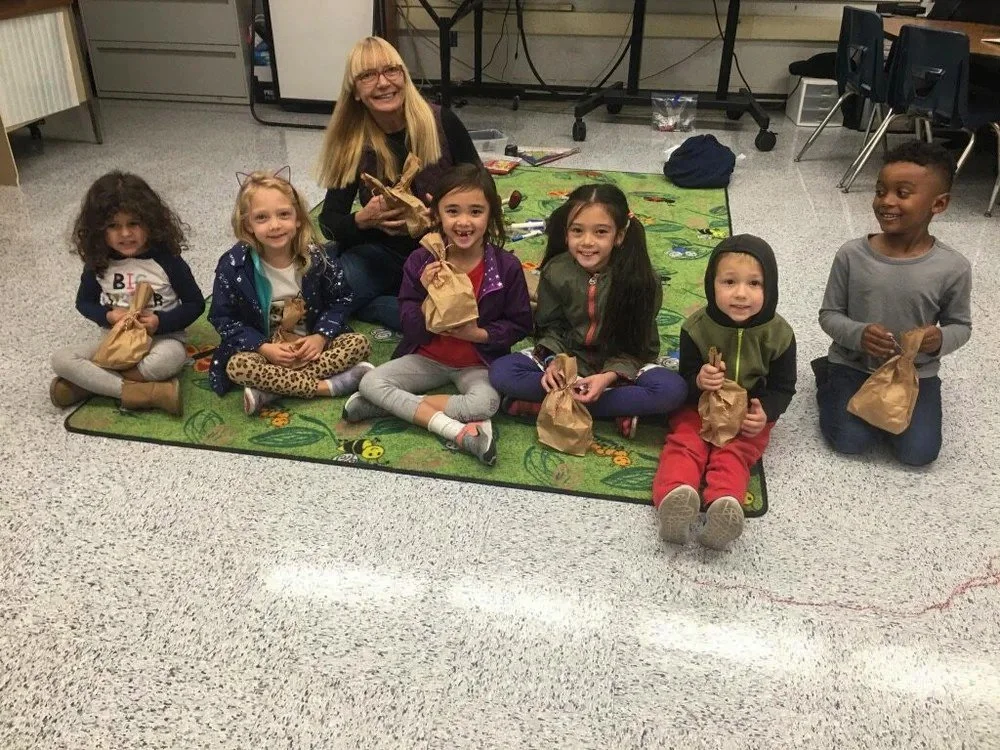 Group of Kinderwerkstatt students and their teacher sitting on a rug indoors, holding brown paper bags.