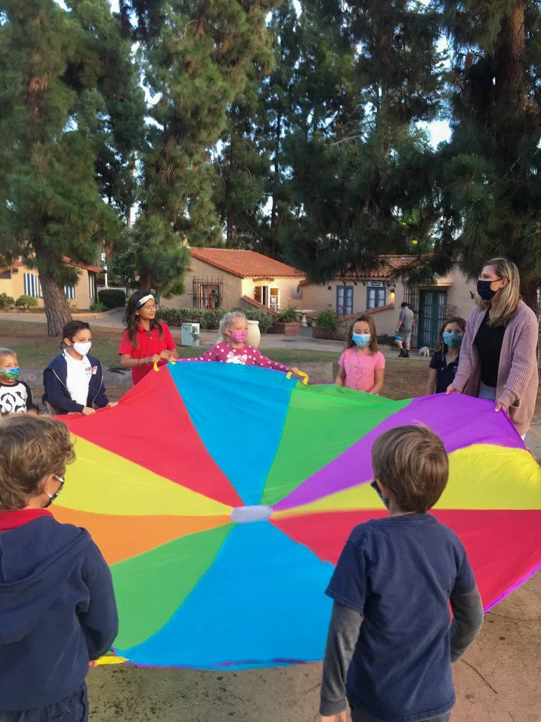 Kinderwerkstatt students playing with a large, colorful parachute in front of the House of Germany cottage during Covid times.