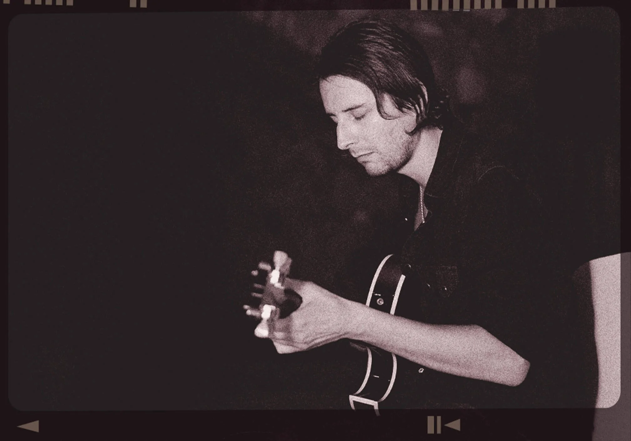 A person with shoulder-length dark hair playing an acoustic guitar with closed eyes, wearing a black shirt, against a dark background.