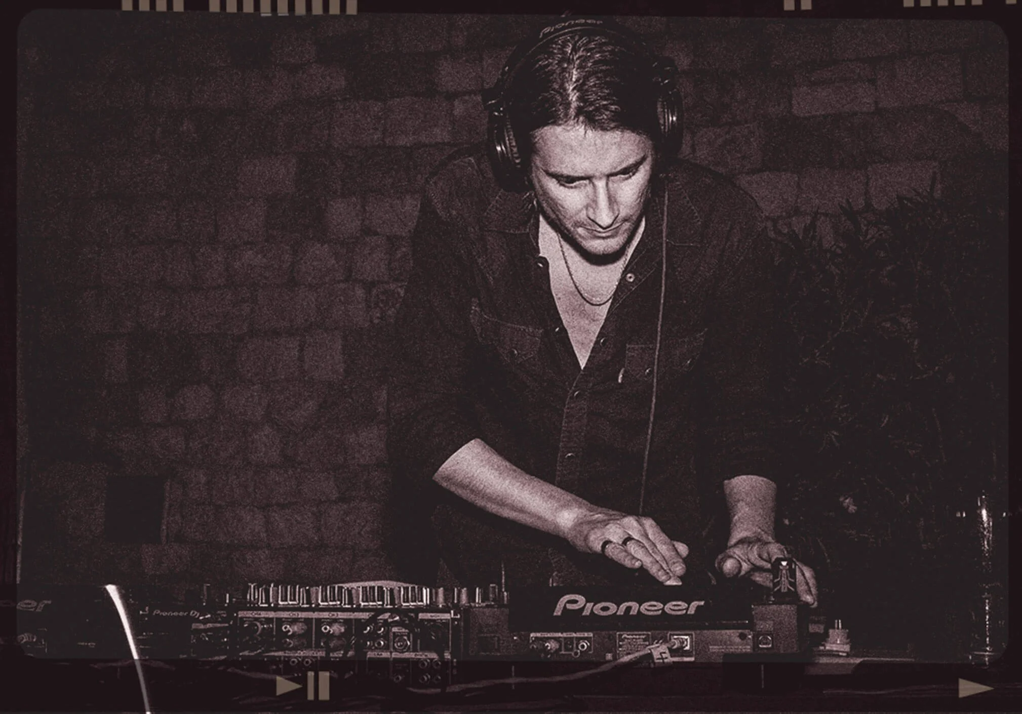 A DJ with headphones using a Pioneer DJ controller at a club or event, with a brick wall background.