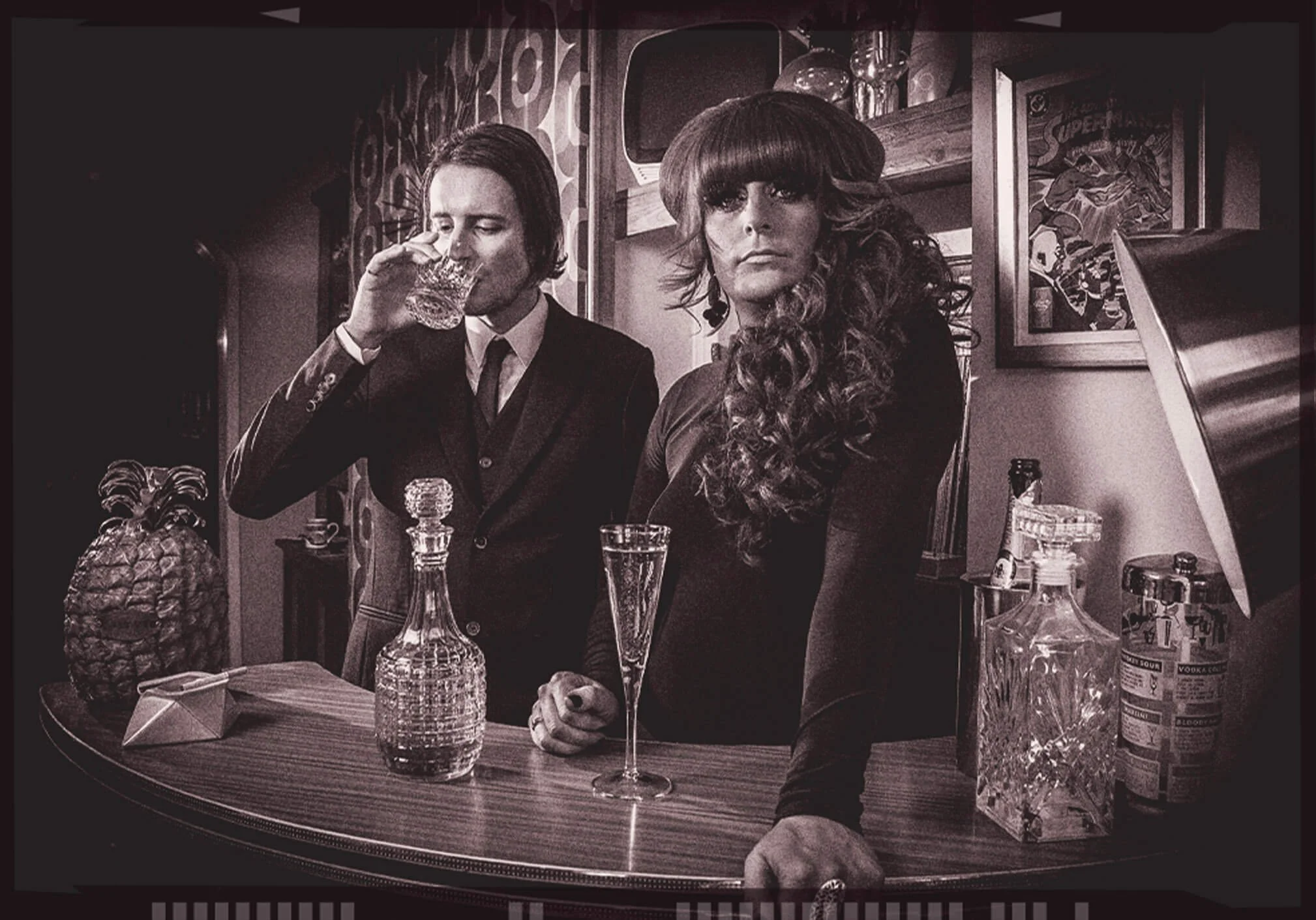 Black and white photo of a man and woman standing behind a bar counter, with drinks and decorations on the counter and shelves behind them, in an indoor setting.