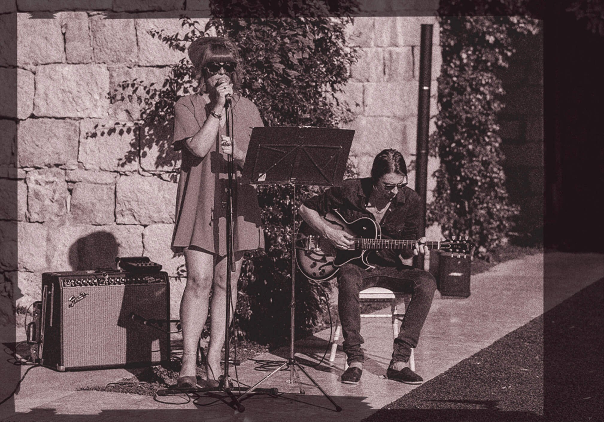 A woman singing into a microphone stands next to a seated woman playing an electric guitar. They are outdoors, against a stone wall with foliage, with a guitar amplifier and music stand nearby.