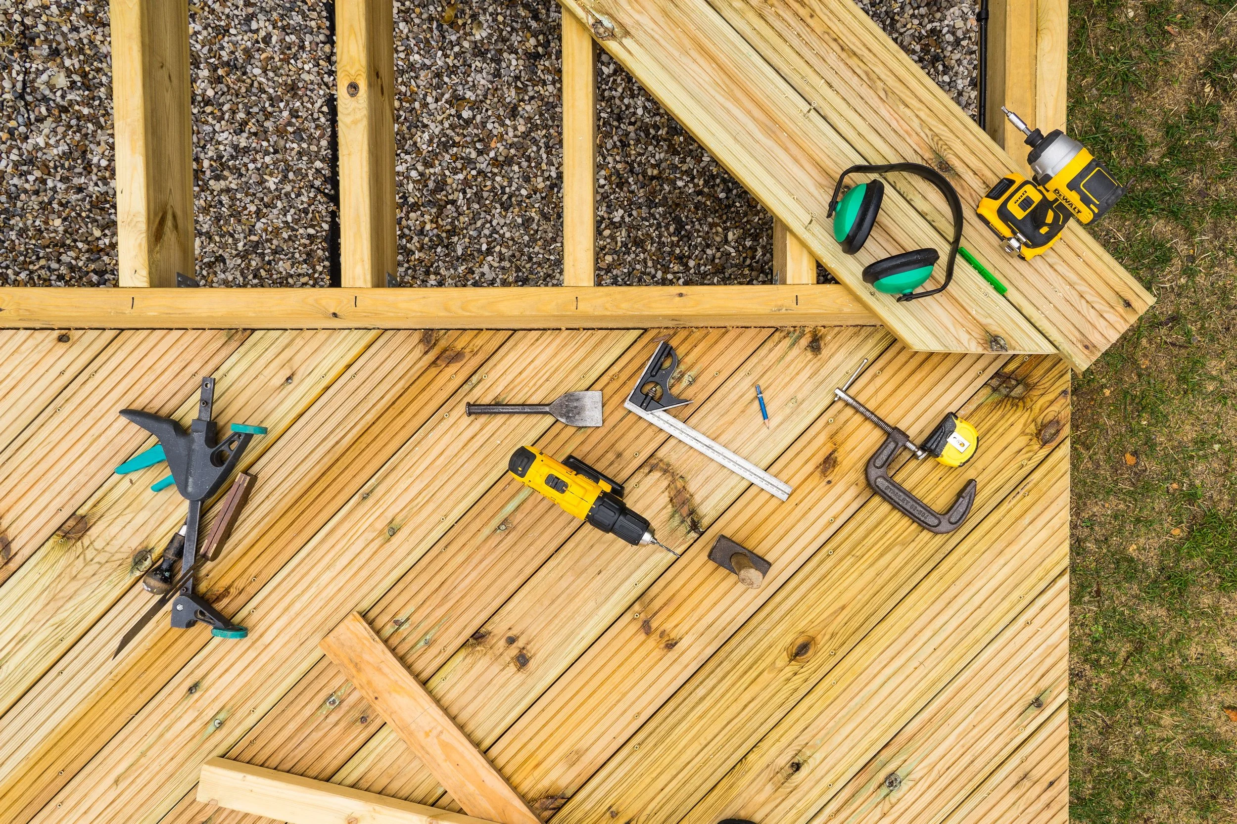 Overhead view of a wooden deck under construction, showing the frame, diagonal floorboards, and professional tools like drills and clamps.