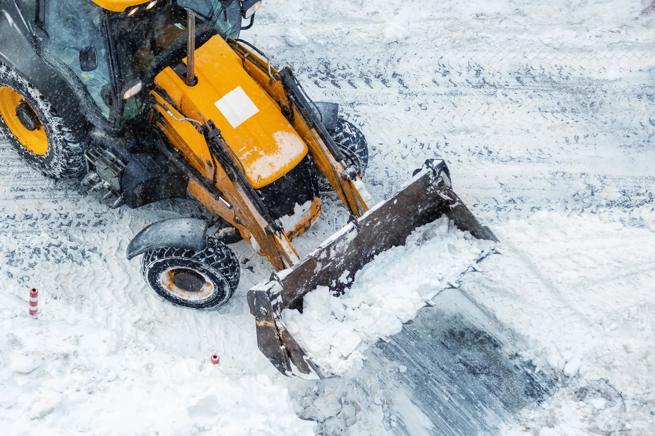 Yellow backhoe loader clearing heavy snow from a paved road using its front bucket during winter maintenance.