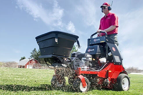 Professional landscaper operating a red stand-on aerator and spreader machine on a large green lawn.