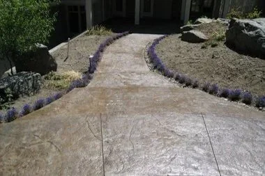 Textured stamped concrete path leading to a home, lined with lavender plants and decorative landscape boulders.
