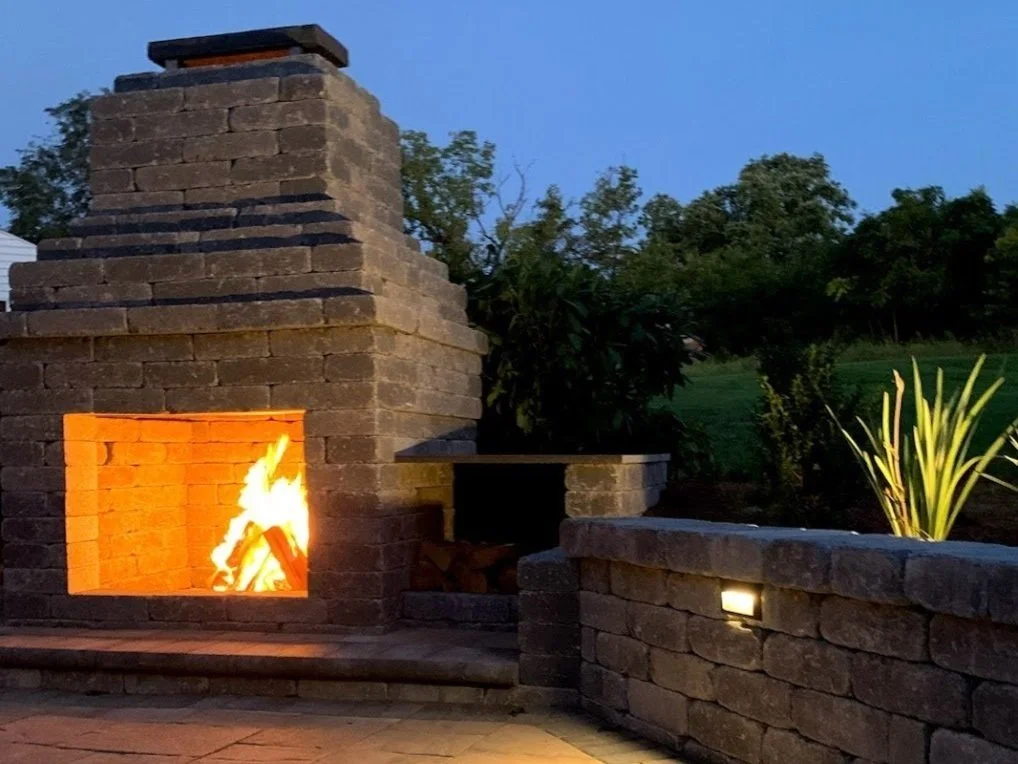 Large stone outdoor fireplace with a roaring fire at night, featuring a lit stone retaining wall.
