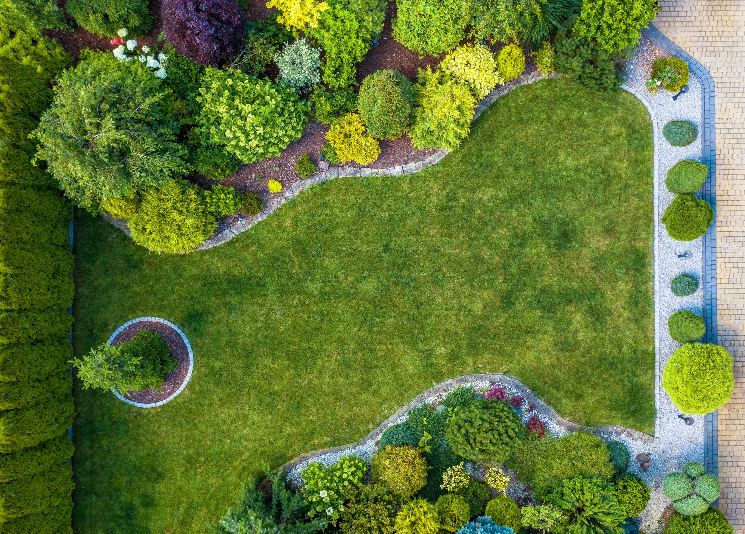 An aerial top-down view of a perfectly manicured lawn surrounded by sculpted garden beds, lush green shrubs, and decorative stone edging.