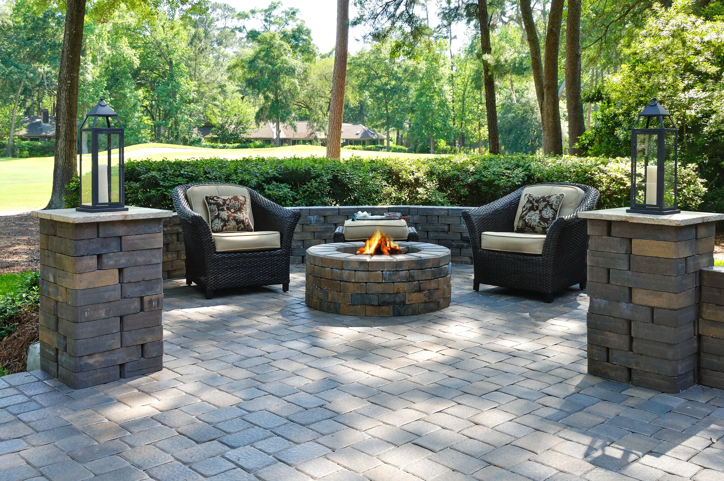 A stone paver patio featuring a central fire pit, wicker armchairs with cushions, and decorative lanterns atop stone pillars.