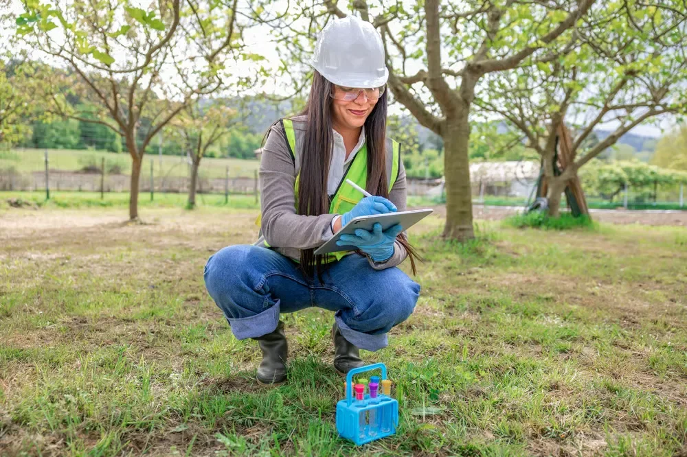 Female lawn care specialist in a hard hat recording soil test data on a tablet in a grove of trees.