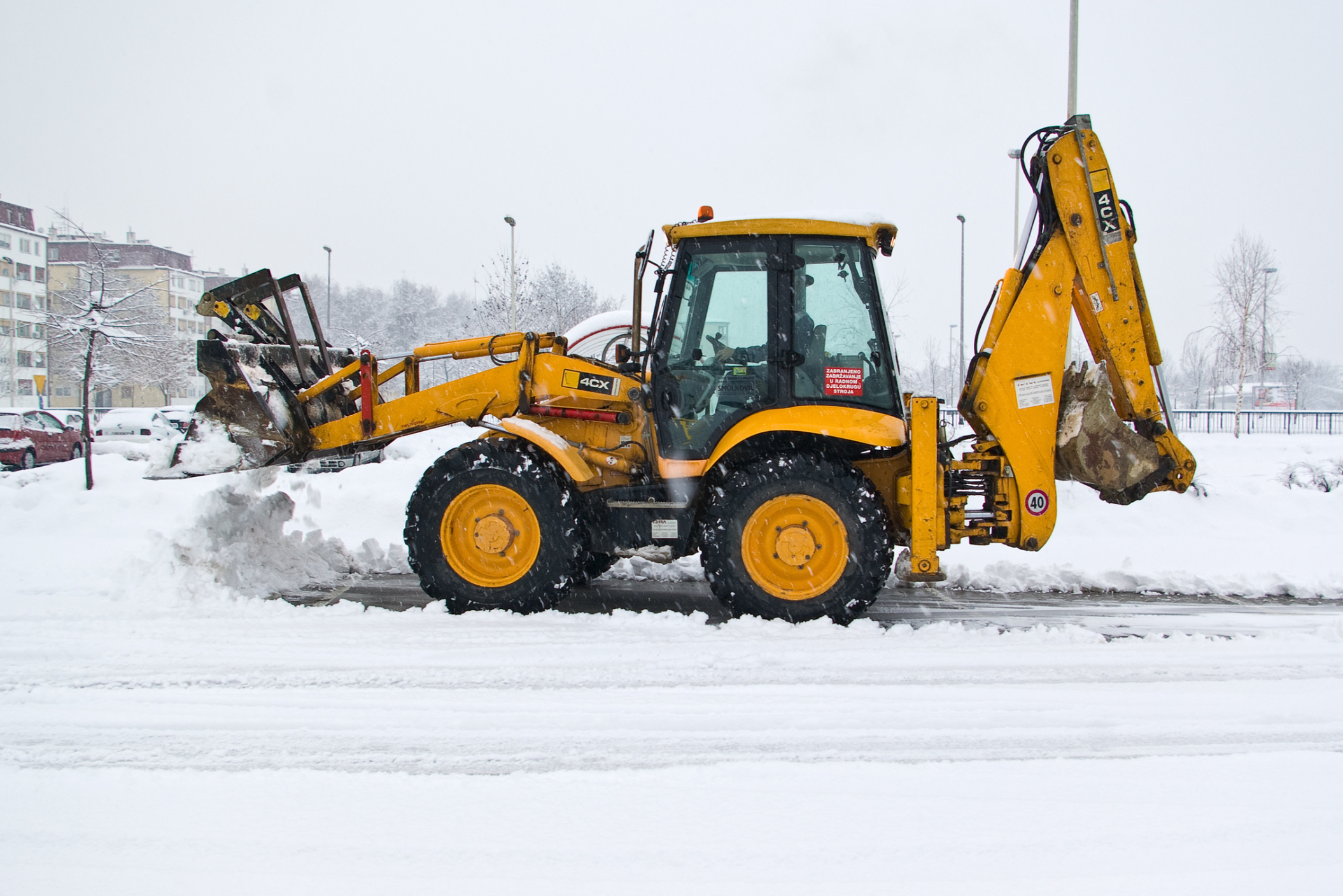 A yellow backhoe loader clears snow from a paved area in a winter city setting.