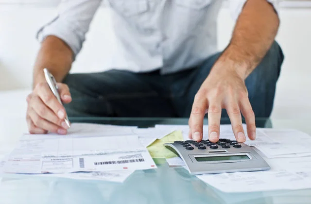 A person using a calculator and holding a pen while reviewing several paper documents.