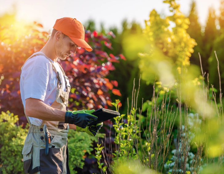 A professional landscaper in an orange cap and work overalls using a digital tablet to manage a garden project outdoors.