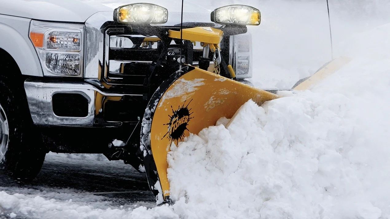 A front-view close-up of a truck with a yellow snow plow clearing deep snow from a road.