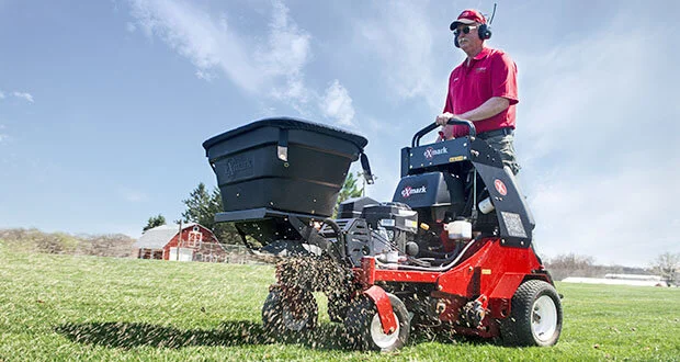 A professional landscaper operating a red ride-on aerator and spreader to treat a large green field under a clear blue sky.